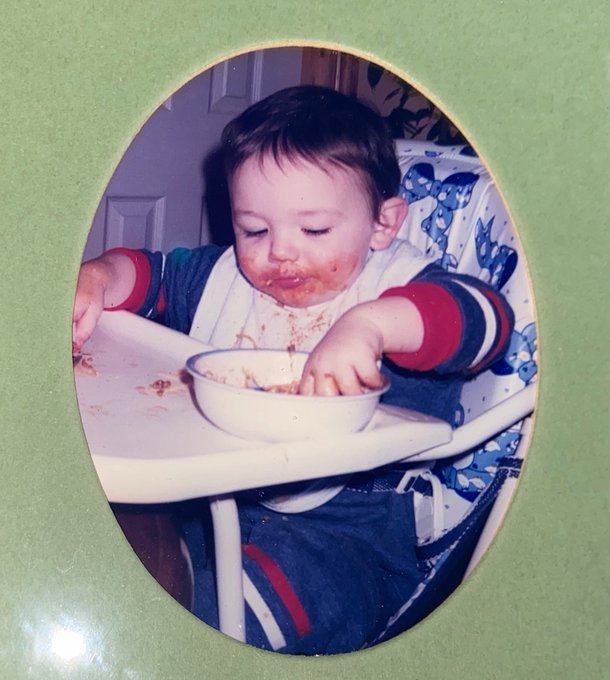 A nostalgic photo of a baby sitting in a high chair, covered in food with a messy face, reaching into a bowl.