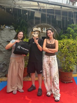 Three people pose together outdoors on a red carpet, holding a banner for 'SÓC CƠM HOÀNG THÀNH' with one carrying a bag labeled 'PrimeHub'.