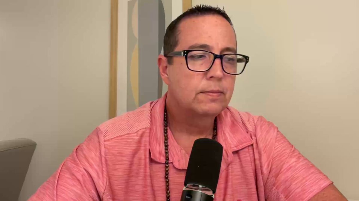 The image shows a man wearing glasses sitting at a desk with a microphone, likely preparing for or just finished recording a podcast or radio show. The background features a white wall that contrasts with the gray carpeted floor beneath him. There are no other people in the scene, and the focus is on the man and his equipment.
