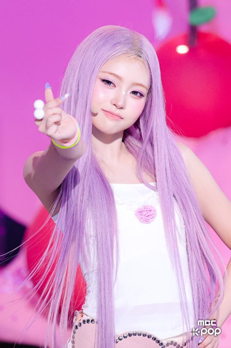 A performer with long, straight lavender hair poses on a brightly lit stage with pink and red backgrounds; she wears a white top with a pink rose detail, and an "MBC k-pop" logo appears in the bottom right corner.