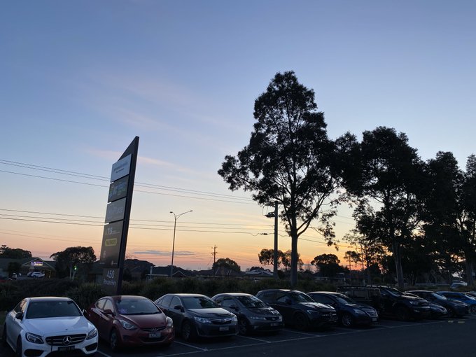 A parking lot at sunset with several parked cars, a tall pylon sign, and silhouetted trees against an orange and blue sky.