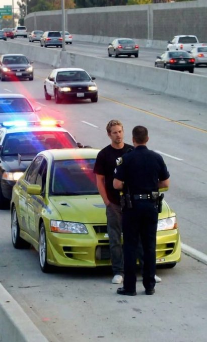 A man in a black t-shirt is being detained by a police officer next to a lime green sports car on a highway shoulder, with a police car with flashing lights visible behind them.
