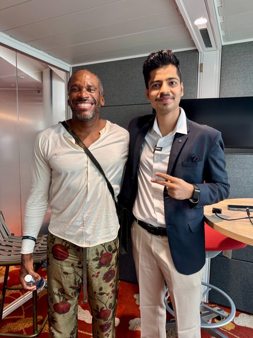 Two men pose together for a photo, one wearing a white shirt and distinctive apple-patterned pants, and the other in a dark suit jacket over a white shirt, smiling in what appears to be a modern office or studio setting.