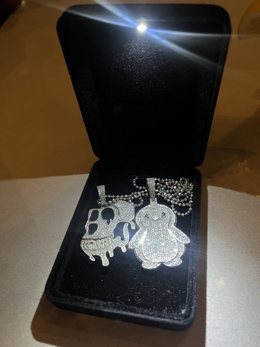 An open black jewelry box displaying two diamond-encrusted pendants shaped like dripping cartoon characters on chains.
