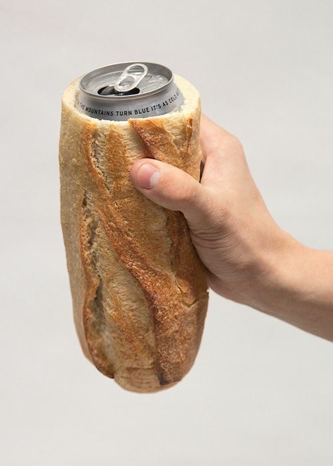 A surreal image of a human hand holding a loaf of crusty bread with a silver soda can protruding from the top, featuring the text 'THE MOUNTAINS TURN BLUE IT'S AS COLD AS...' on the can.