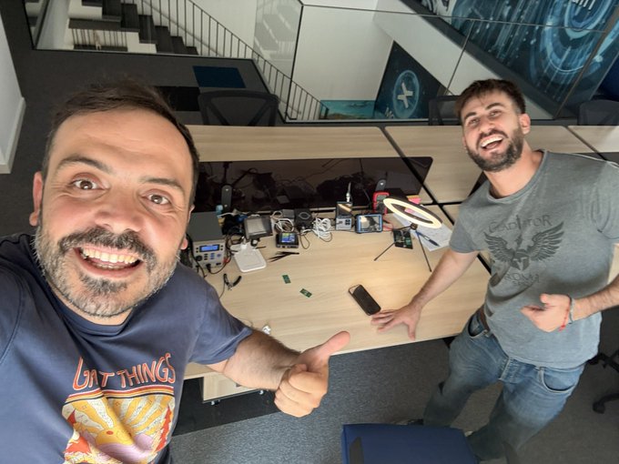 Two men smile and give thumbs up in a tech workshop or lab, with one wearing a 'WATTINGS' t-shirt and the other wearing a grey shirt, surrounded by electronic equipment on a workbench.