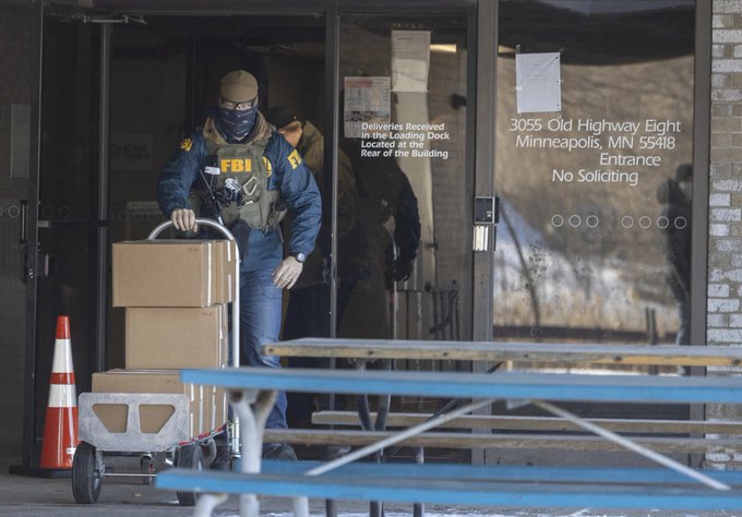 An FBI agent wearing a face covering and tactical vest exits a building at '3055 Old Highway Eight, Minneapolis, MN 55418', pushing a hand truck loaded with cardboard boxes labeled as evidence.