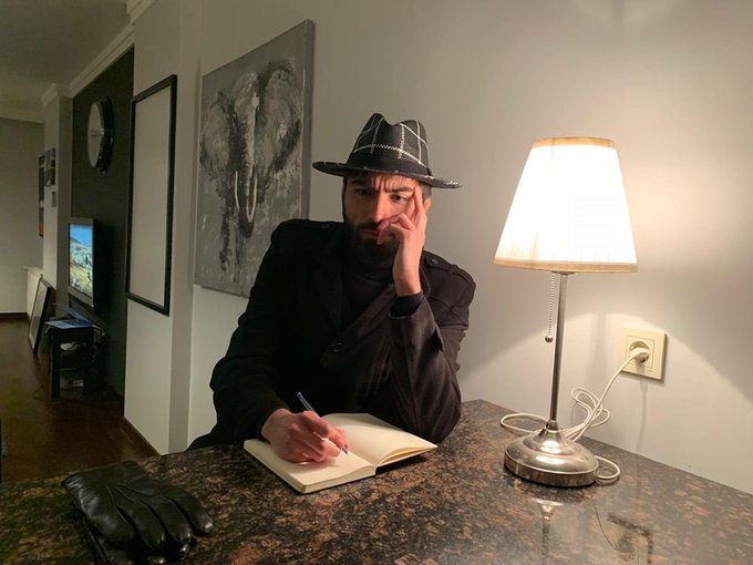 A bearded man wearing a black hat and dark jacket sits at a desk writing in a notebook, illuminated by a table lamp.