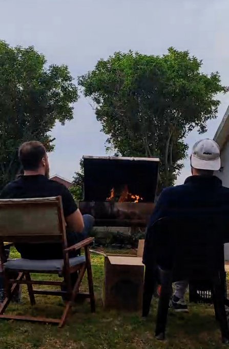 Two men sit in a backyard facing a black grill with an active fire, surrounded by trees under an overcast sky.