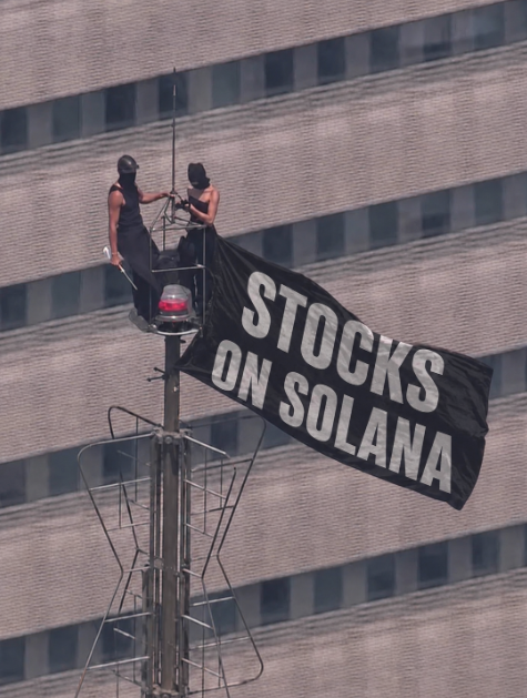 Two people standing on top of a tall structure against a skyscraper, unfurling a black flag that reads 'STOCKS ON SOLANA'.
