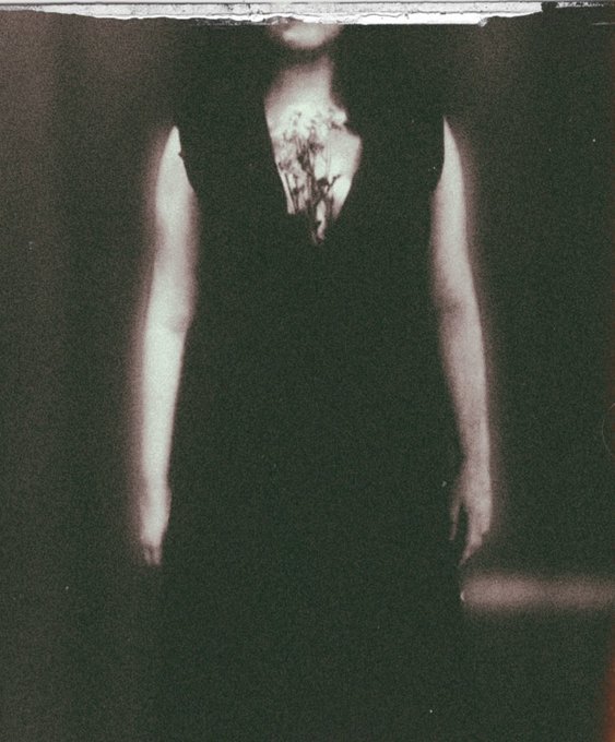 A dark, blurry photograph shows a figure in a black top with white floral patterns across the chest, arms extended outward against a nearly black background, with what appears to be a cylindrical object (possibly a microphone) visible in the lower right corner.

The image thematically aligns with the post's message of "blooming through the ache"—the floral pattern blooming from the figure's chest visually echoes the metaphor of growing flowers from pain, embodying the resilience described in the accompanying text about NFT piece 2/5 on the XTZ (Tezos) blockchain.