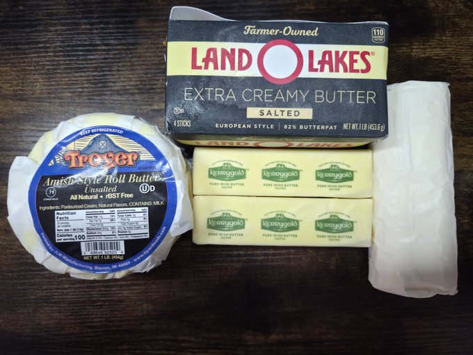 An assortment of butter products on a wooden table, including a Treyer Amish-Style Roll Butter, a Land O'Lakes Extra Creamy Butter box, and Kg Saaggold butter sticks.