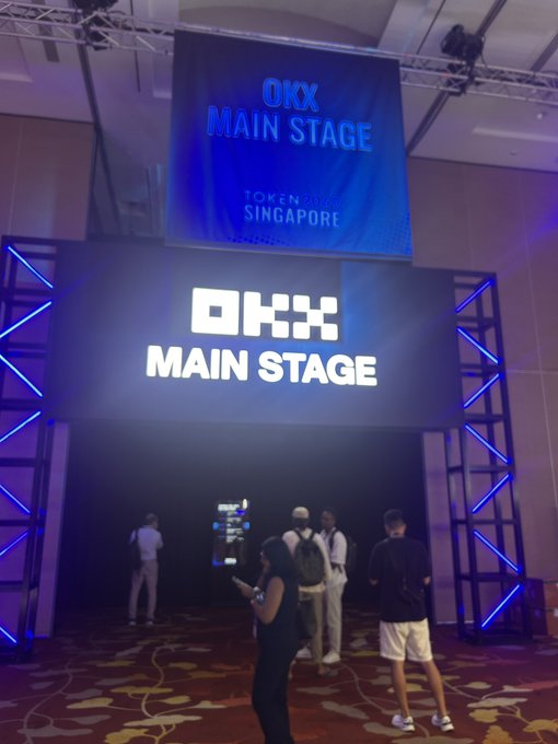 A photograph of the OKX Main Stage entrance at the Token2049 Singapore conference, featuring large illuminated signage with people walking through the doorway.