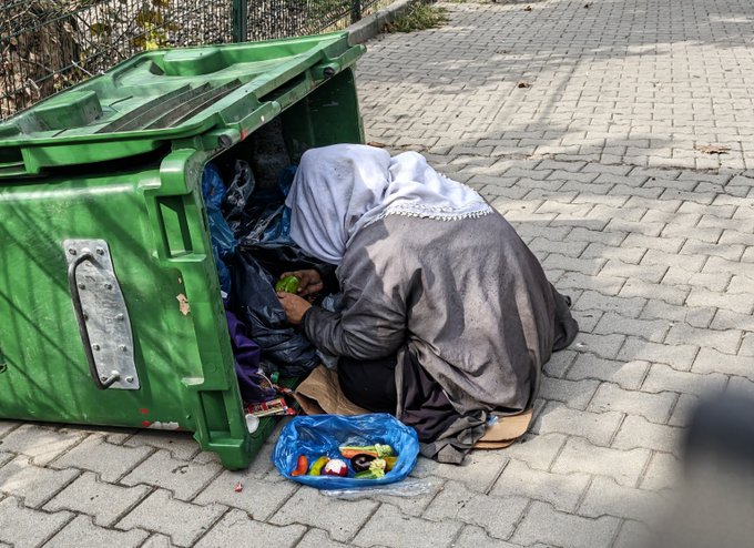 A person wearing a grey jacket and light blue headscarf crouches on a paved street, rummaging through an open green dumpster with a blue plastic bag nearby.