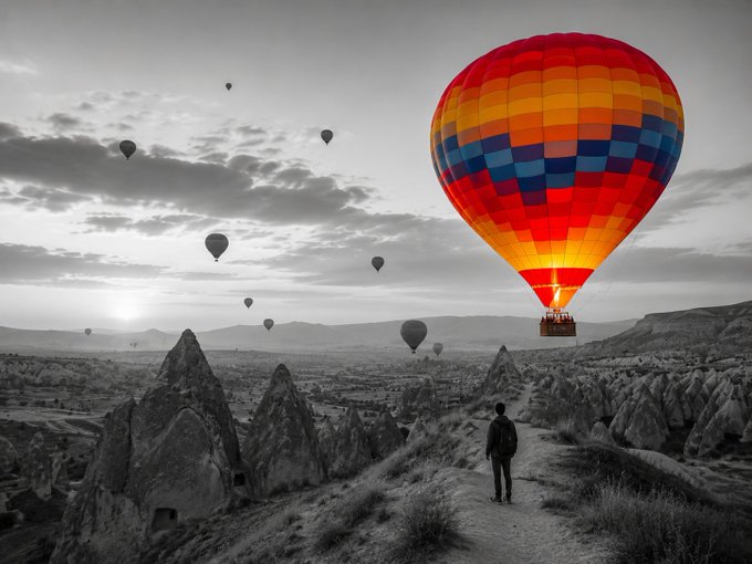 A black and white photograph of a person standing on a rocky path in Cappadocia, watching multiple hot air balloons, with one balloon highlighted in vibrant color.