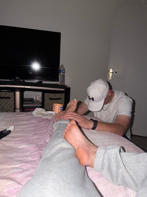 A man wearing a white baseball cap and t-shirt massages the feet of a person lying on a bed in a dimly lit room.