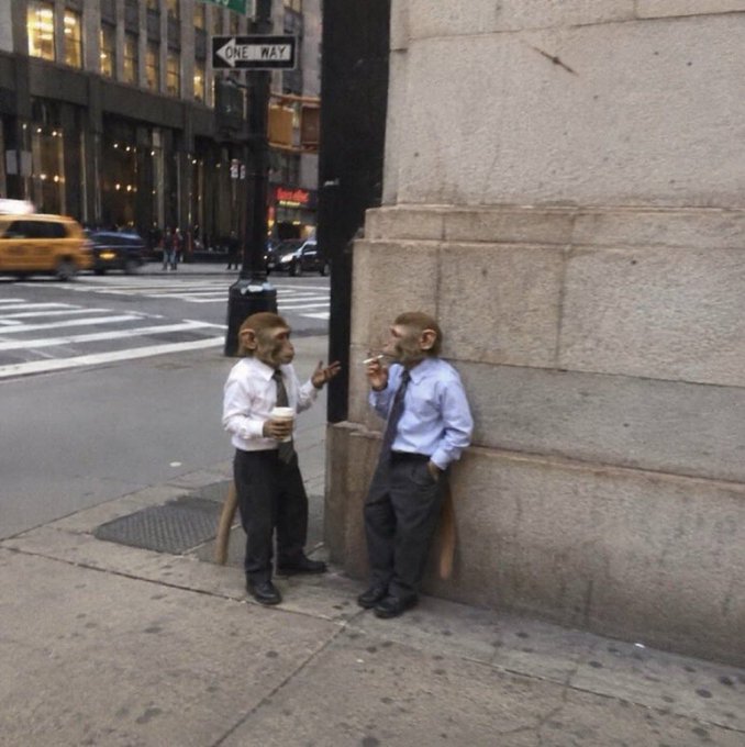 Two people wearing monkey masks and business attire (white shirt with tie, blue shirt with tie) stand on a city street corner having a smoke break next to a building, with a "ONE WAY" sign and yellow taxi visible in the background.