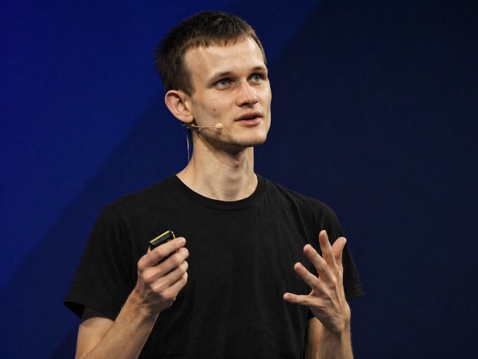 Vitalik Buterin speaks at a conference, wearing a black t-shirt and headset microphone, gesturing with his hands while holding a presentation clicker. The image accompanies news of his proposed "Lean Ethereum" initiative, a multi-year overhaul of the Ethereum protocol.