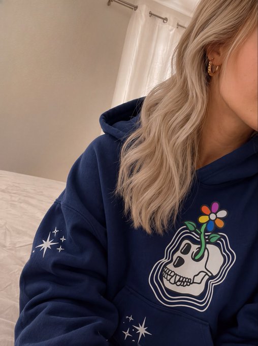 A selfie-style photo of a person with wavy blonde hair wearing a navy blue hoodie featuring a graphic of a skull with a colorful flower growing from it and small star designs.