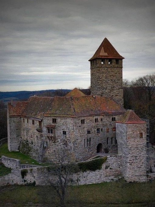 Photograph of an old stone castle or fortress with a tall square tower and red-tiled roofs, set against a cloudy sky with hills visible in the distance.