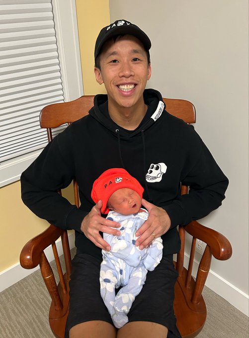 A smiling man wearing a black cap and hoodie sits in a wooden rocking chair holding a sleeping newborn baby dressed in a blue cloud-patterned onesie and red beanie.