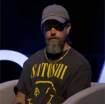 A man wearing a cap and sunglasses, dressed in a t-shirt with 'SATOSHI' and a Bitcoin logo printed in yellow.