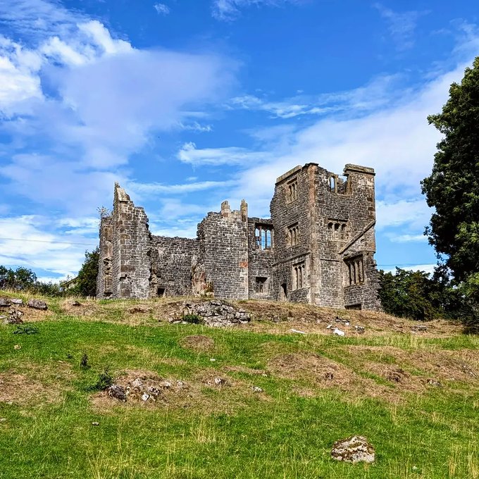 The stone ruins of a castle or large building stand on a grassy hill under a bright blue sky with scattered clouds.