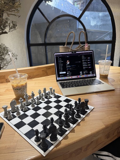 A wooden table holds a chessboard set up for a game, alongside an iced coffee, a glass drink, and a laptop displaying a Discord chat.