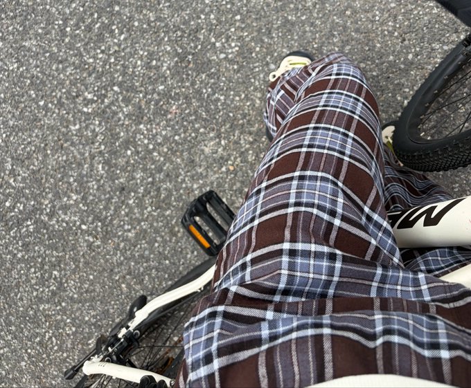 A person in brown plaid pants straddles a white bike (with visible "M" lettering on the frame) on gray pavement, apparently gearing up to ride. The image reflects the post's humorous take on choosing cycling as a money-losing alternative to inactivity.