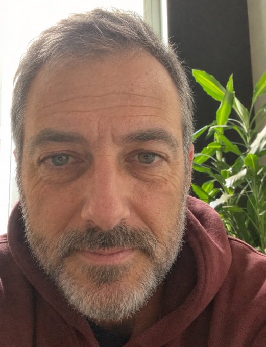 A close-up selfie of a middle-aged man with short graying hair, blue eyes, and a gray beard, wearing a maroon hoodie, with green houseplants visible in the background.