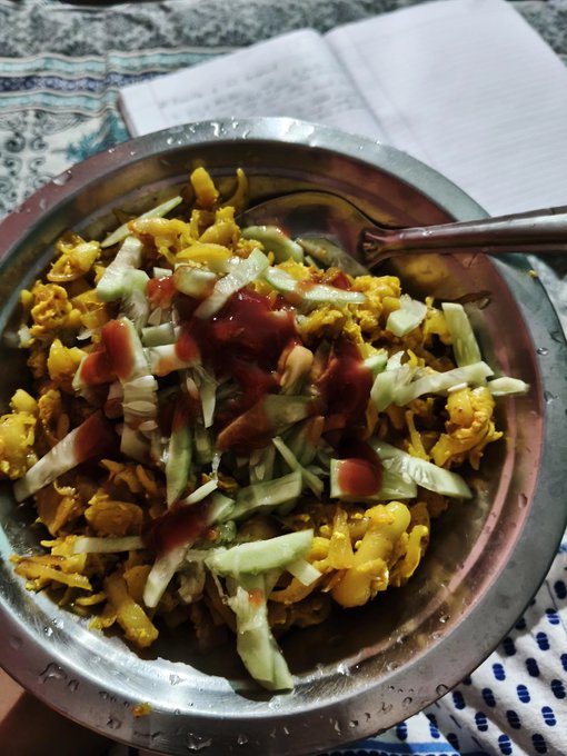A metal bowl containing a meal of scrambled eggs topped with shredded cucumbers and drizzled with red sauce, served with a spoon on a patterned cloth.