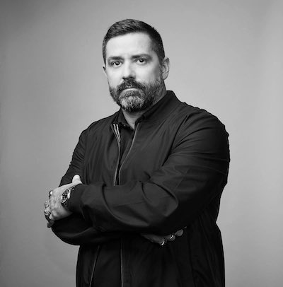 A black-and-white professional portrait of a bearded man wearing a dark jacket, standing with his arms crossed and wearing a silver wristwatch against a plain gray backdrop.