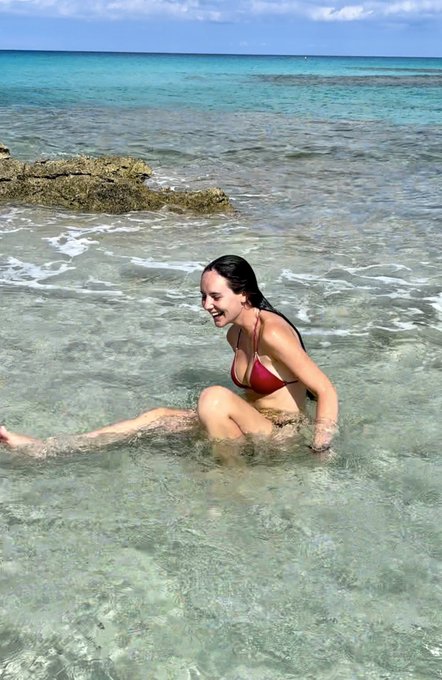A young woman in a red bikini sits smiling in shallow, crystal-clear turquoise water at a tropical beach, with rocky outcrops visible in the background under a partly cloudy sky.