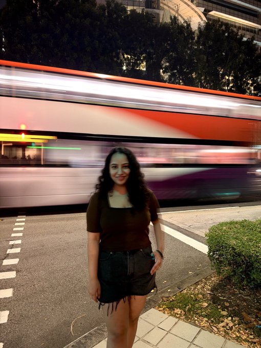 A young woman in a brown top and dark shorts stands on a city street at night, smiling as a blurred red and white bus passes behind her.