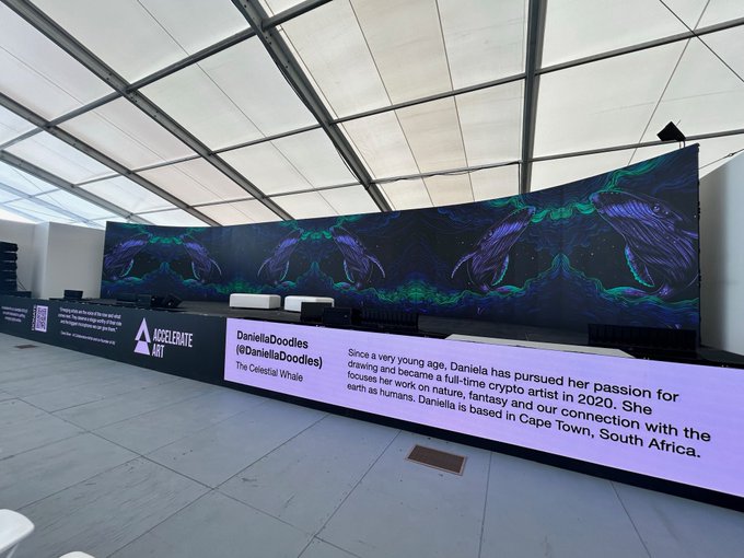 The image shows a large screen displaying an advertisement for an art exhibit called "The Celestial Whale". The text on the screen reads, "Good morning and happy Friday! Remembering when 'The Celestial Whale' was on display at Miami Art Basel with @accelerateart still available on @SuperRare 0.25 ETH buy now."