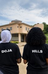 Two people stand with their backs to the camera wearing shirts labeled "DOJA" and "HILLS" in front of a building, marking their successful in-person outreach at the GenLayer Agent Tank Hackathon where they onboarded new participants with cupcakes.