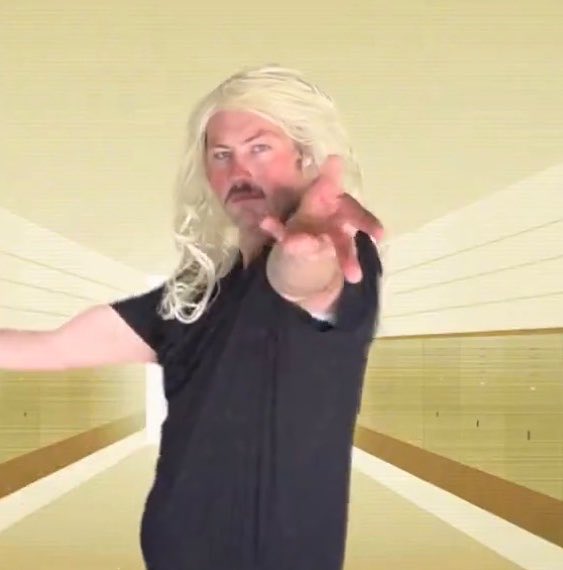 A man with a blonde wig and mustache points dramatically at the camera while wearing a black t-shirt against a sunburst background.