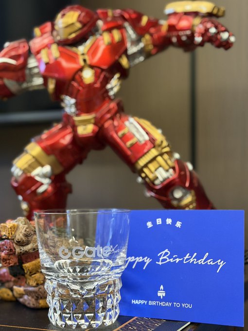 The image features a glass of water with "Happy Birthday" written on a blue card placed next to it, along with a statue of Iron Man. The scene suggests that someone has prepared a special birthday celebration for an Iron Man fan or a person who enjoys the character from the Marvel Cinematic Universe.