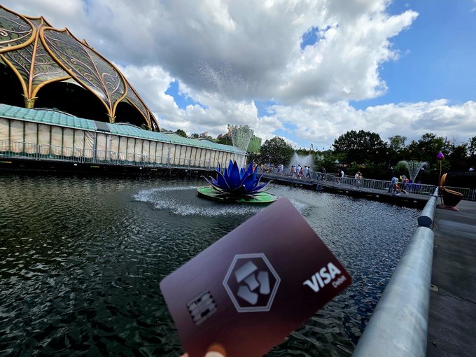 A hand holding a dark crypto-themed Visa debit card in front of a pond with fountains and a decorative blue lotus sculpture at what appears to be a theme park.