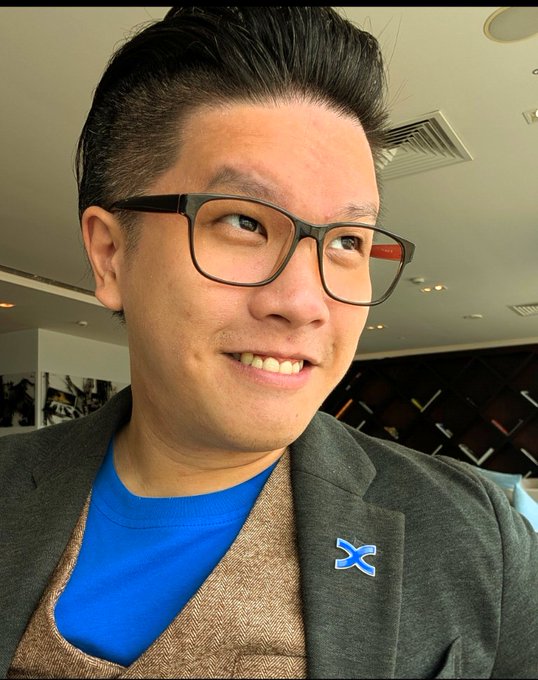A man with dark hair and black glasses smiles while wearing a blue shirt, brown vest, and gray blazer with a blue X pin.