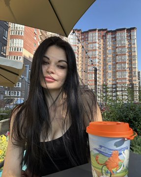 A selfie of a young woman with long dark hair wearing a black top, sitting outdoors at a café with a fruit-patterned drink cup beside her.