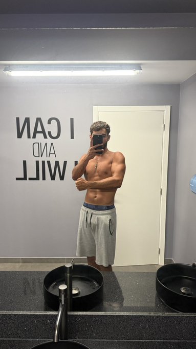 A shirtless man in gray sweatpants takes a mirror selfie in a gym restroom; mirrored text on the wall reads "I CAN AND WILL".