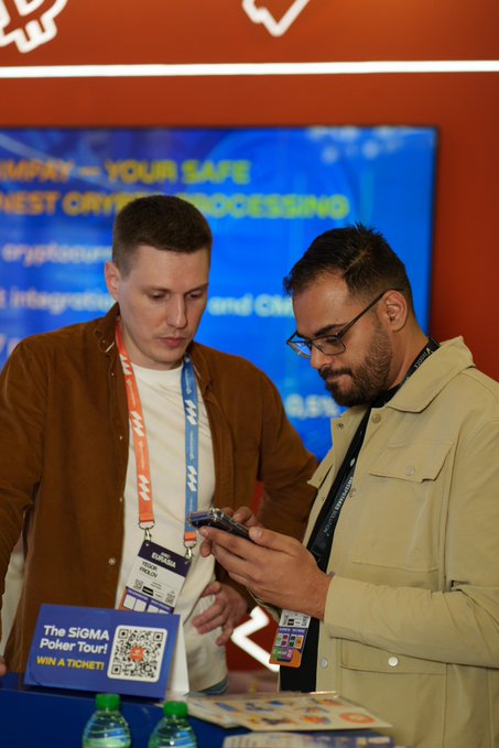 Two men wearing conference lanyards look down at a smartphone together at what appears to be a crypto event, with a SIGMA Poker Tour QR code sign visible in the foreground.