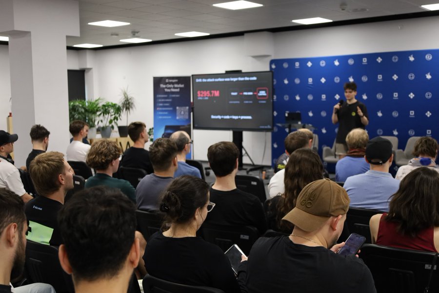 An audience attends a presentation in a conference room, where a speaker stands next to a large screen displaying data about crypto wallets and a $295.7M figure.
