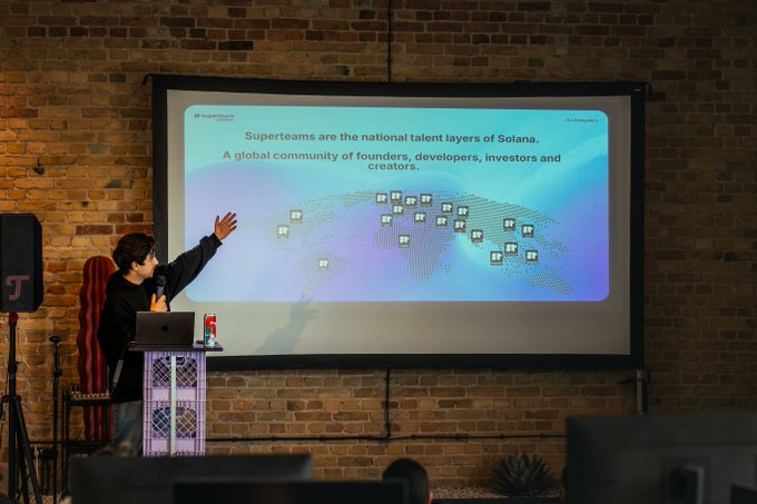 A speaker at a podium presenting in front of a large screen that displays a world map made of dots and the text 'Superteams are the national talent layers of Solana. A global community of founders, developers, investors and creators.'