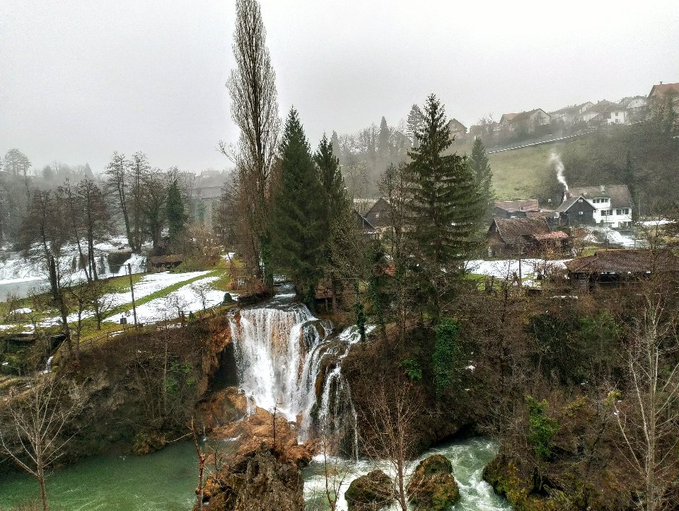 A serene waterfall cascades through a misty, snow-dusted village landscape with evergreen trees and traditional houses, evoking the tranquil escape that awaits after the relentless grind of chart-watching and crypto min-maxing.