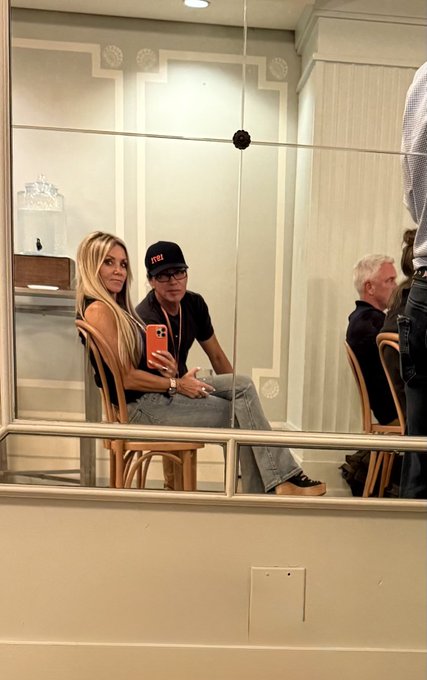 A stylish blonde woman in jeans and a black top takes a selfie with a bearded man wearing a "Proof" baseball cap and glasses inside an elegant venue, fitting the post's playful caption referencing crypto terms—him as "Proof of Money" and her as "Proof of Honey." The man also wears a gold chain with what appears to be a Bitcoin or crypto-related pendant, reinforcing the Bitcoin Alpha context.