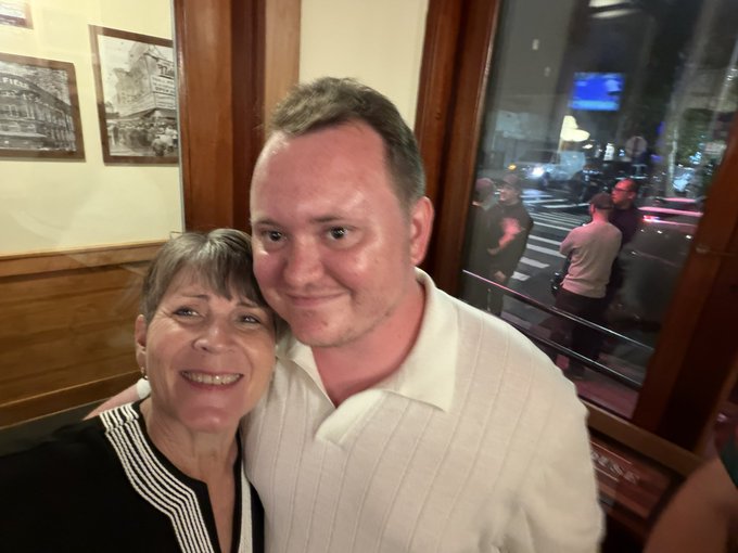 A selfie of an older woman with short gray hair smiling alongside a younger man in a white collared shirt, taken inside what appears to be a restaurant with framed pictures and a window showing a street at night.