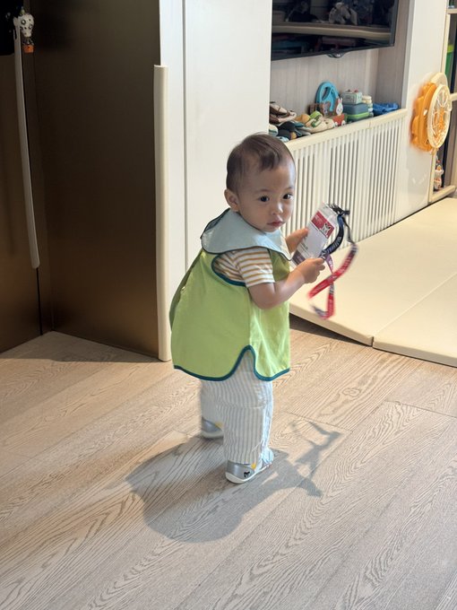 A toddler in a green bib and striped shirt stands on a wooden floor, holding a small plastic object and looking toward the camera.
