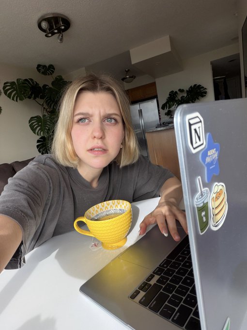 A young woman with a confused expression sits at a white desk with a yellow mug and a laptop covered in stickers, including 'N' and 'HERE COMES TROUBLE'.